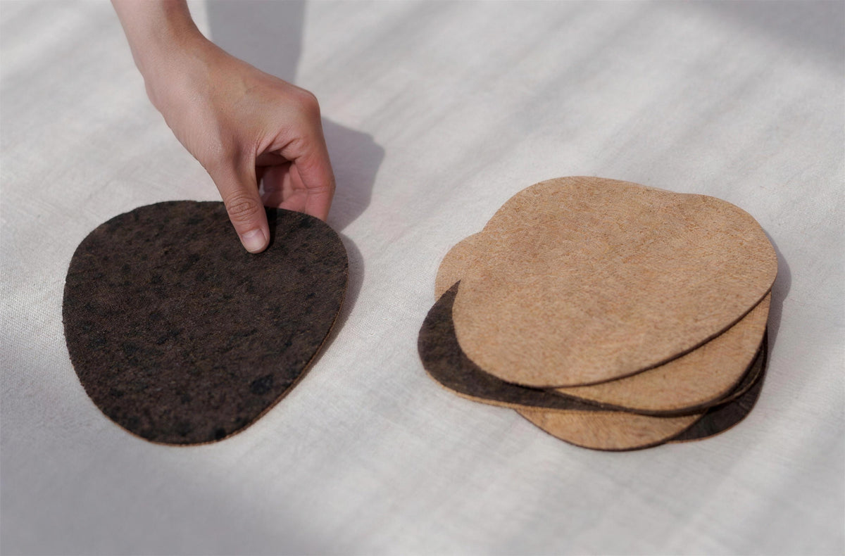 Eine Hand, die neben einem Stapel aus Kork-Tischsets ein dunkles Kork-Tischset aufhebt